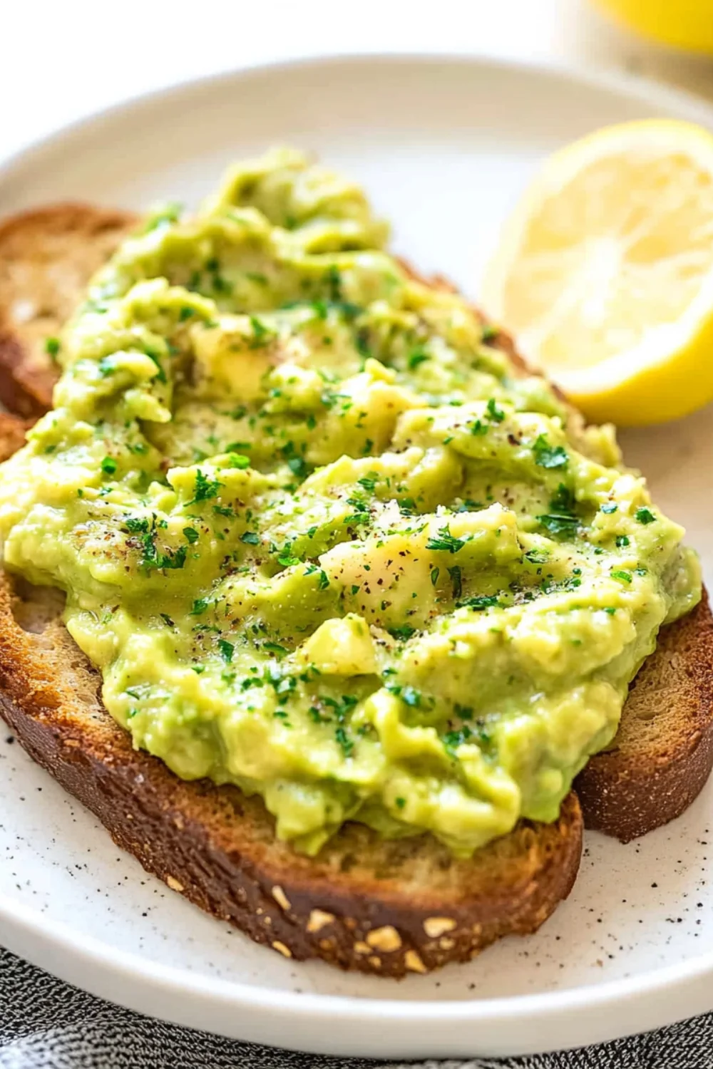 Crispy whole-grain toast topped with creamy mashed avocado, sprinkled with fresh herbs and cracked black pepper, served with a lemon wedge.
