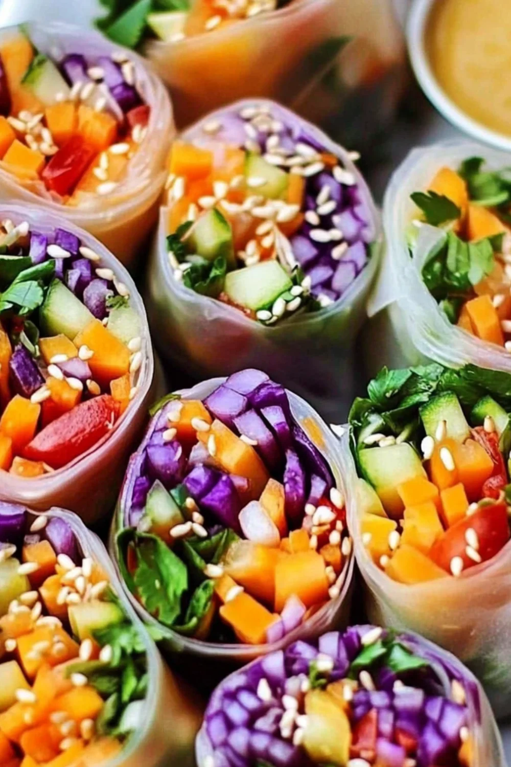 A close-up of Rainbow Veggie Spring Rolls filled with colorful vegetables like purple cabbage, carrots, cucumber, and fresh herbs, topped with sesame seeds and served with a dipping sauce.